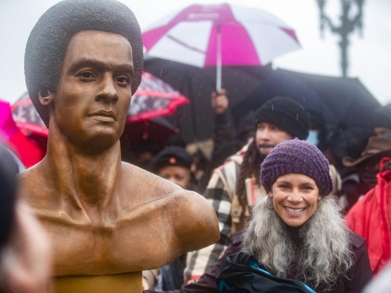 a large shining brown and gold sculpture of a Black man next to petite African American woman who is smiling wearing a purple crocheted hat and flowing curly long grey hair