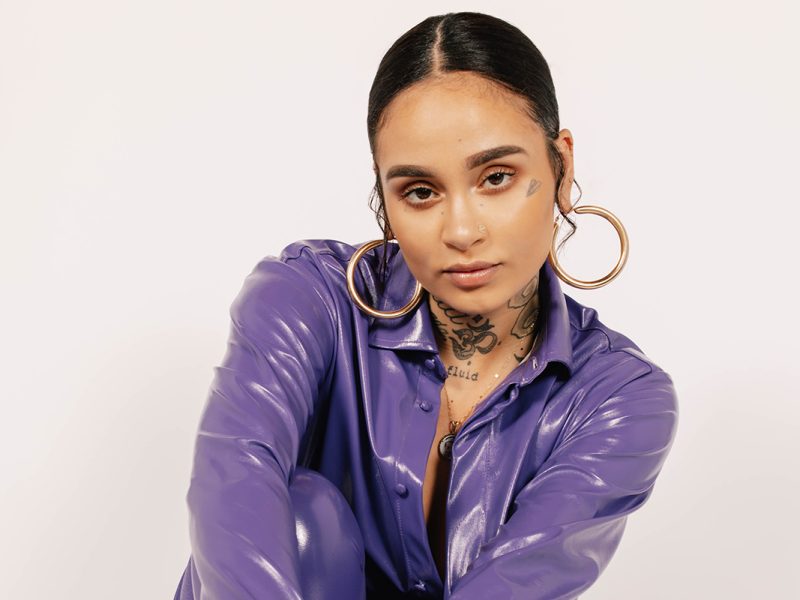 A biracial woman with slicked back hair and tattoos on her face is wearing a purple vinyl jacket