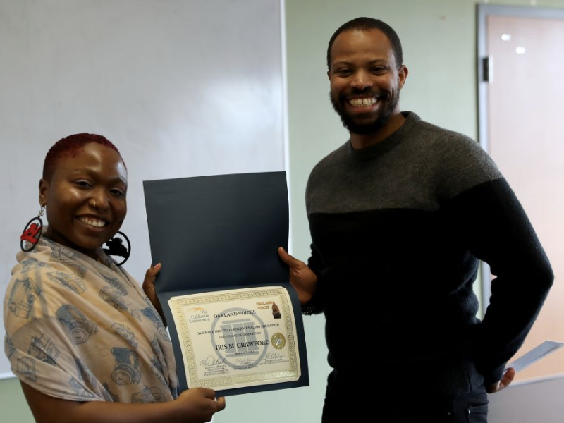 An African American woman with short hair holds a diploma with a taller Black man and they are both smiling.