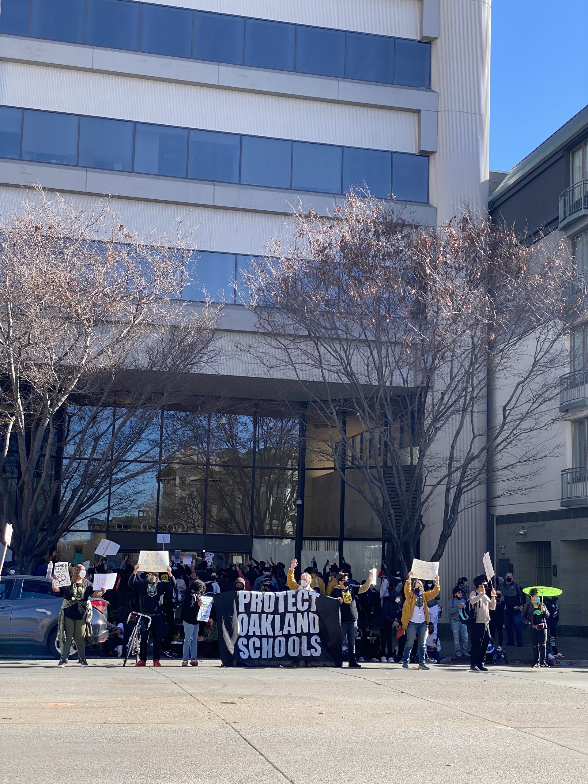 a large group holds a sign facing traffic "Protest Oakland Schools"