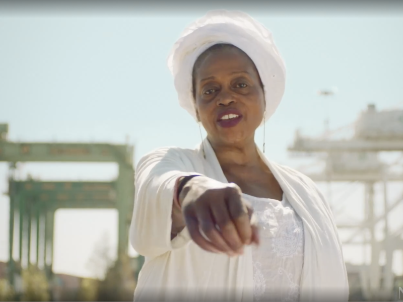 An African American woman wearing all white and a white hair wrap rolls dice towards camera. In the background is Oakland's cranes and shipyards.