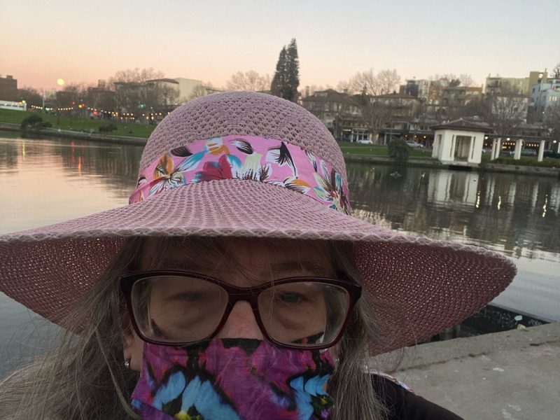 A woman wearing a purple mask, pink hat, and glasses takes a selfie in front of Lake Merritt.