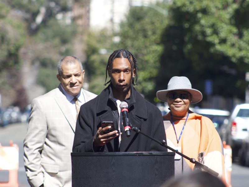 A Black man in an off white suit, a young Black man wearing a black jacket, and Black woman with sunglasses, smiling, in grey hat stand outside in front of podium