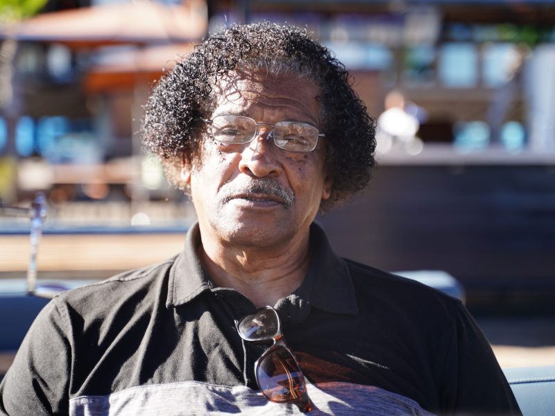An African American man with curly hair and glasses sits outside on a sunny day