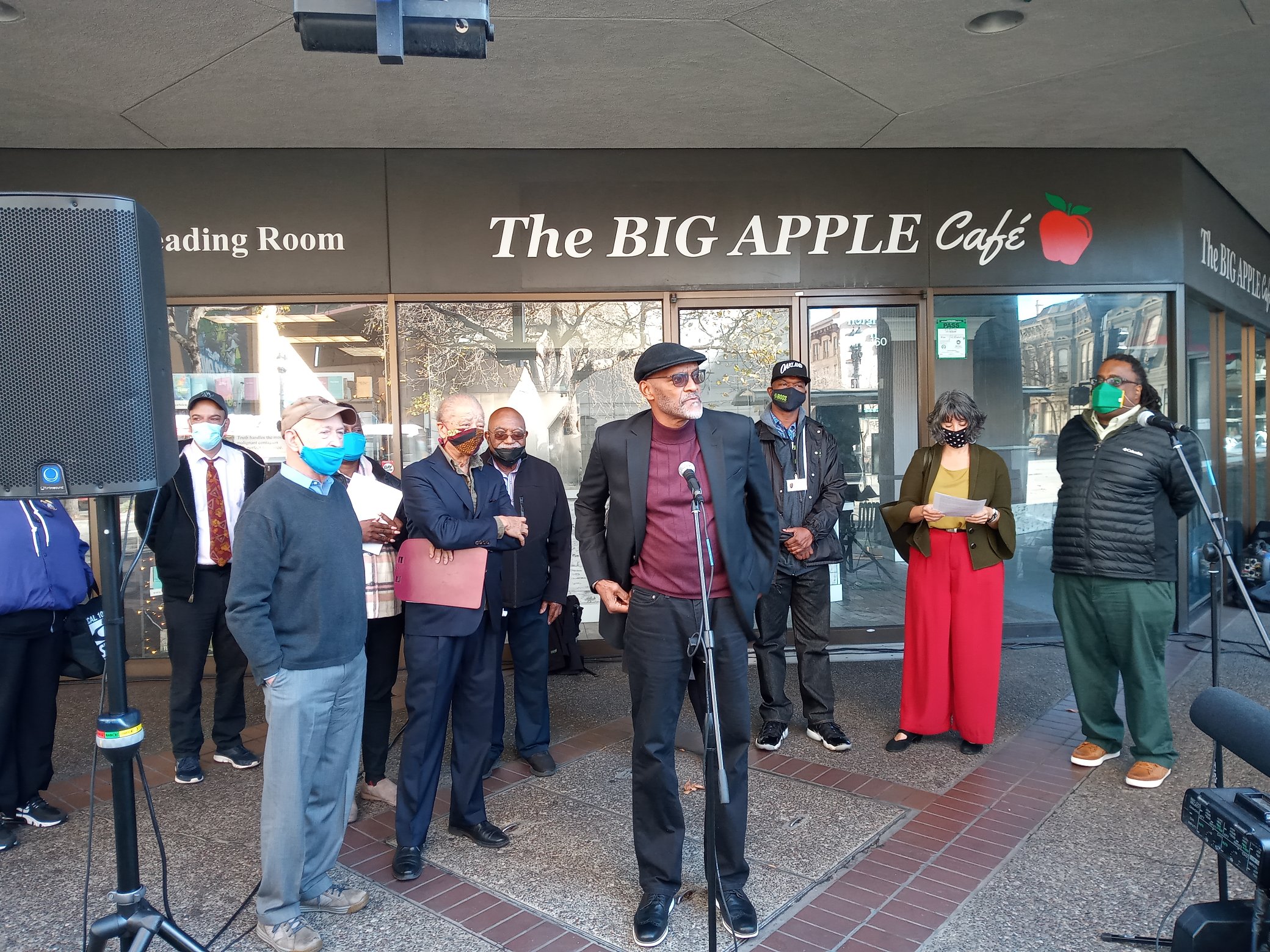 A tall African American man stands in front of "The Big Apple" cafe in downtown Oakland speaking into a mic