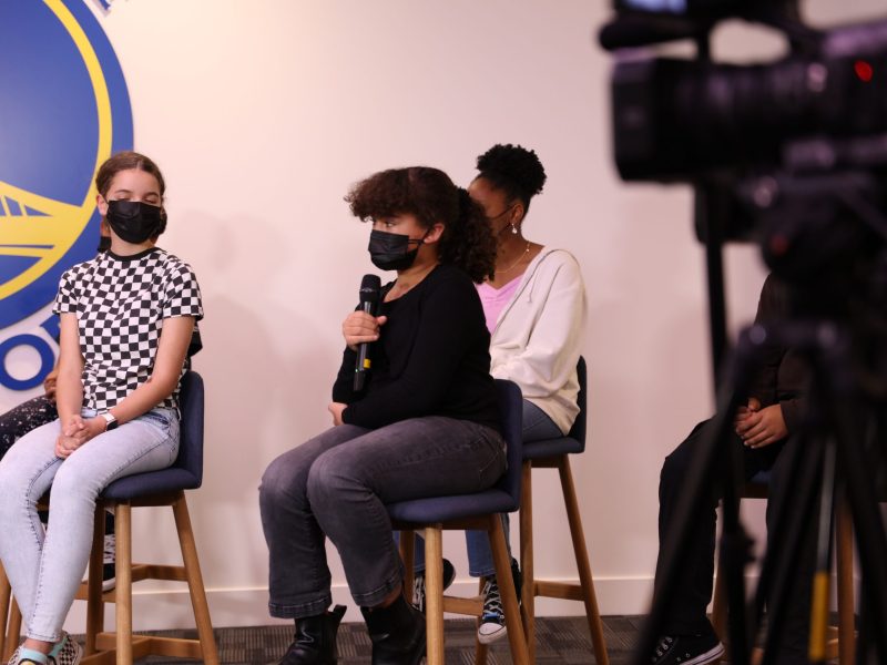 An African American girl wearing a masks speaks into a mic while another students is behind her in front of a Golden State Warriors sign