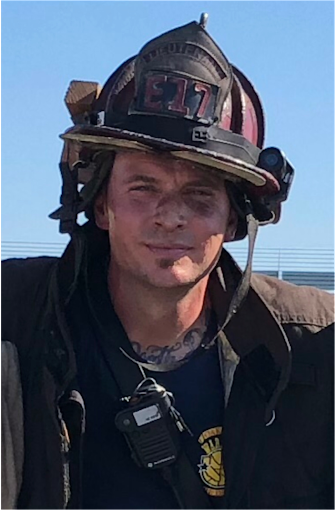 A man wears a firefighters hat with soot covering his face