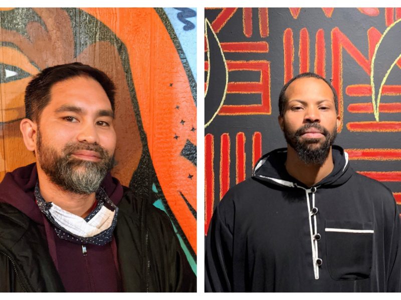 A Filipino American man with grey beard in front of orange mural; an African American man with beard and short hair in black hoodie in front of red and black mural