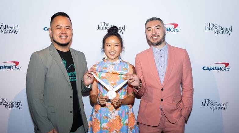 A Filipino American man, Chinese American woman, and Chinese American man dressed in festive outfits stand in front of backdrop that says "James Beard"