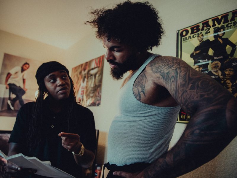 An African American director with a beanie chats with a tall man in a white tank top and with an Afro
