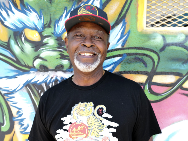An elder African American man wears Warriors cap and black t-shirt with a lion/dragon image on it, standing in front of bright mural.