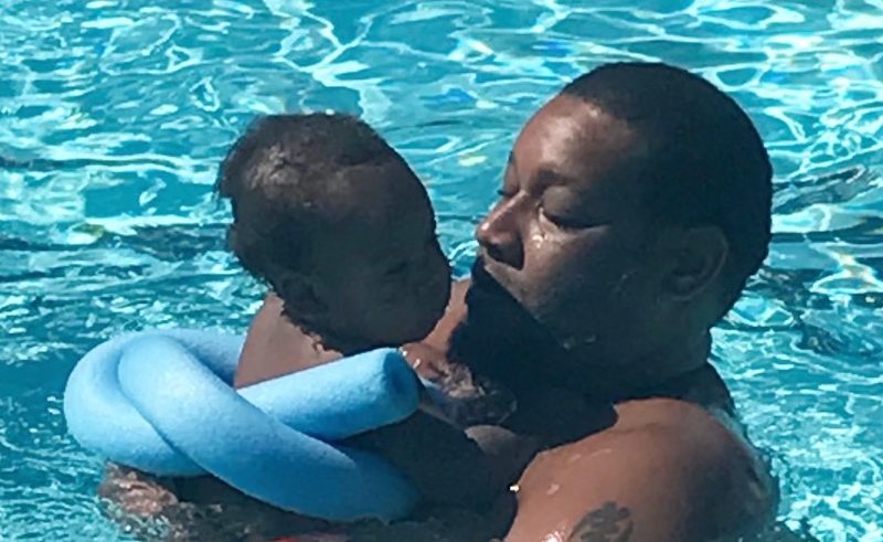 A Black man holds his infant son in the water with a blue pool noodle tied safely around the baby to keep him afloat
