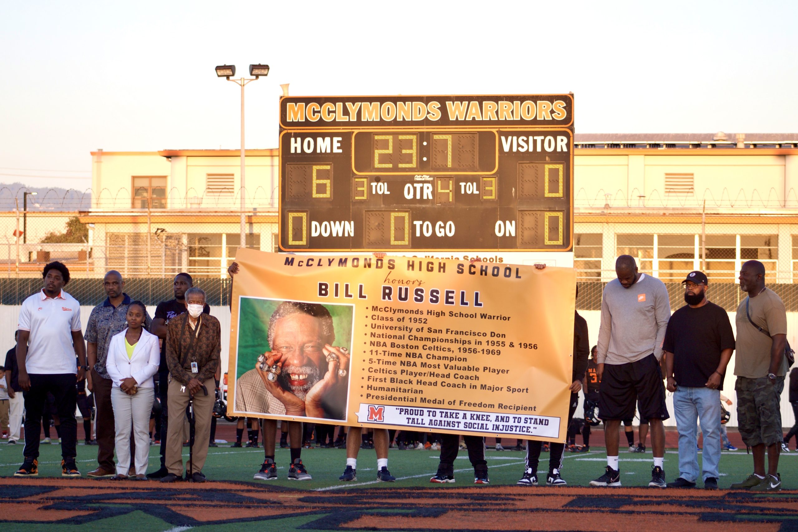 A group of African American men and one woman stand next to banner honoring NBA great Bill Russell on a football field