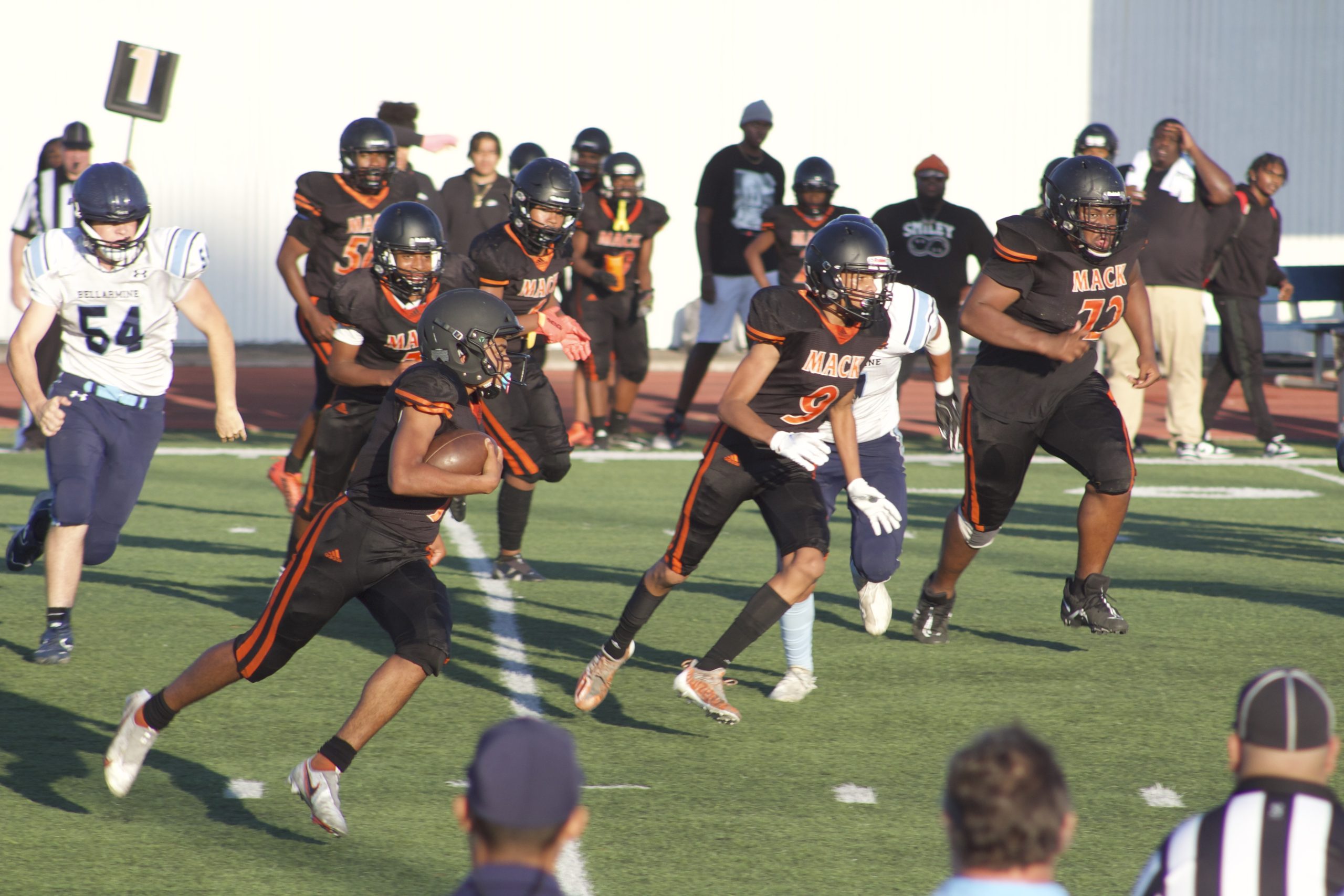 A group of boys playing in a jv football game, with one running hugging football towards a touchdown.