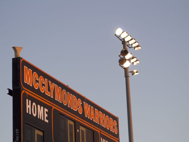 a close up of a scoreboard that says "McClymonds Warriors" in orange type. A field lamp post is shining at dusk next to it