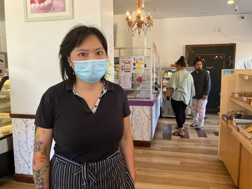 An Asian woman with short hair, face mask, and colorful tattoos on her arm stands in front of a small bakery