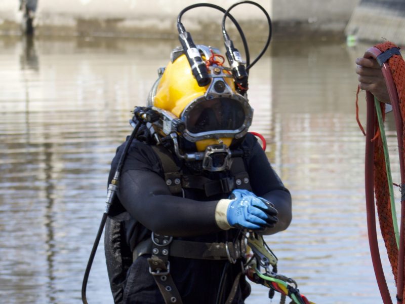 A man in a wetsuit and a yellow hard helmet comes out of murky waters