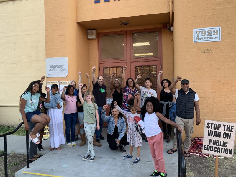 A group of mostly Black students and a few adults stand in front of peach colored school building with sign that says "stop the war on public education"