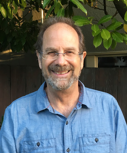 An older white man with speckled beard and glasses smiles for camera