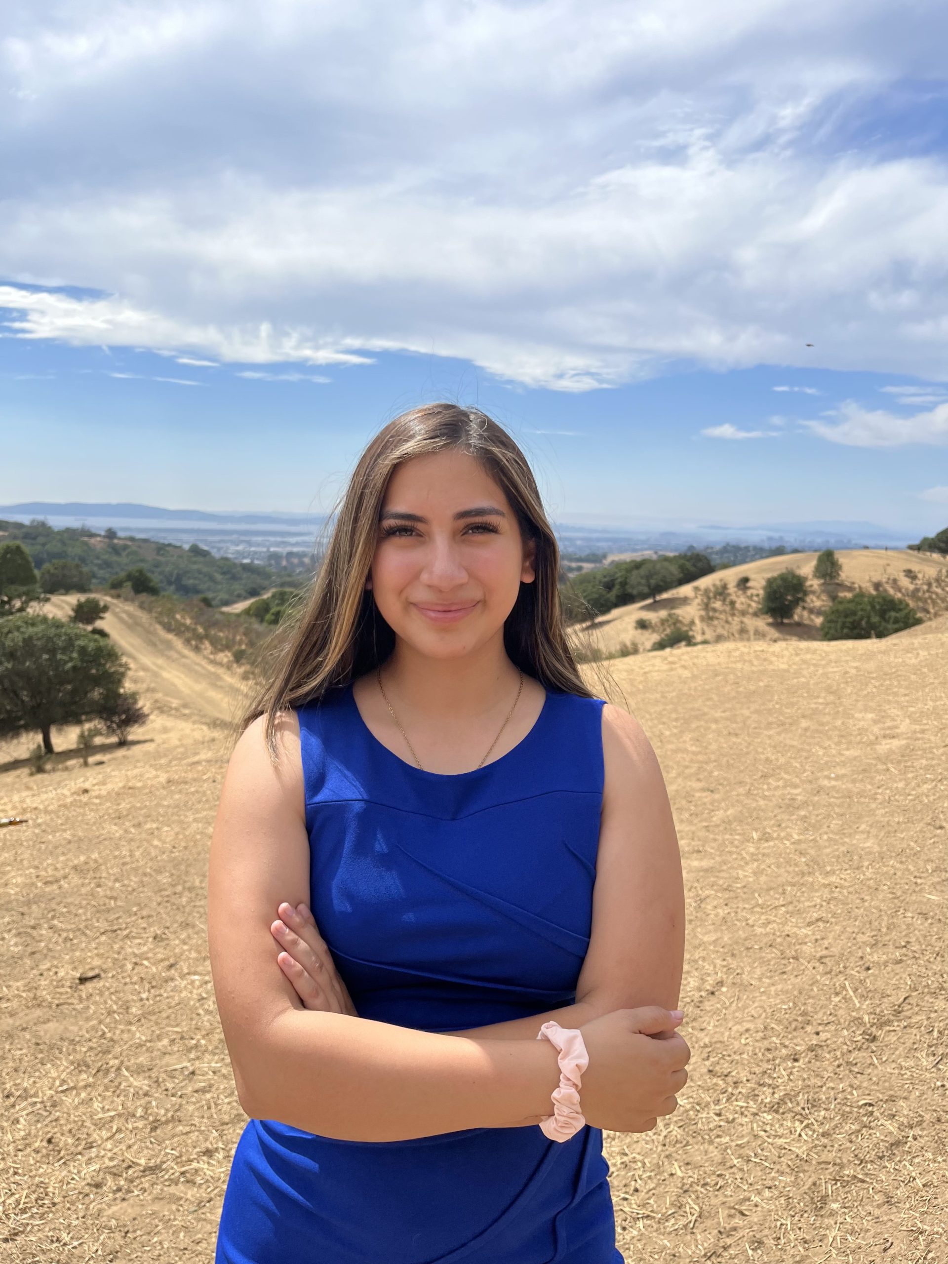 A Latina young woman with blue dress and straight hair stands outside in sun
