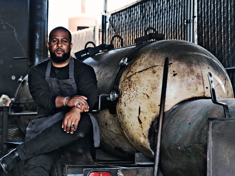 A Black man sits next to a barbecue smoker wearing an apron
