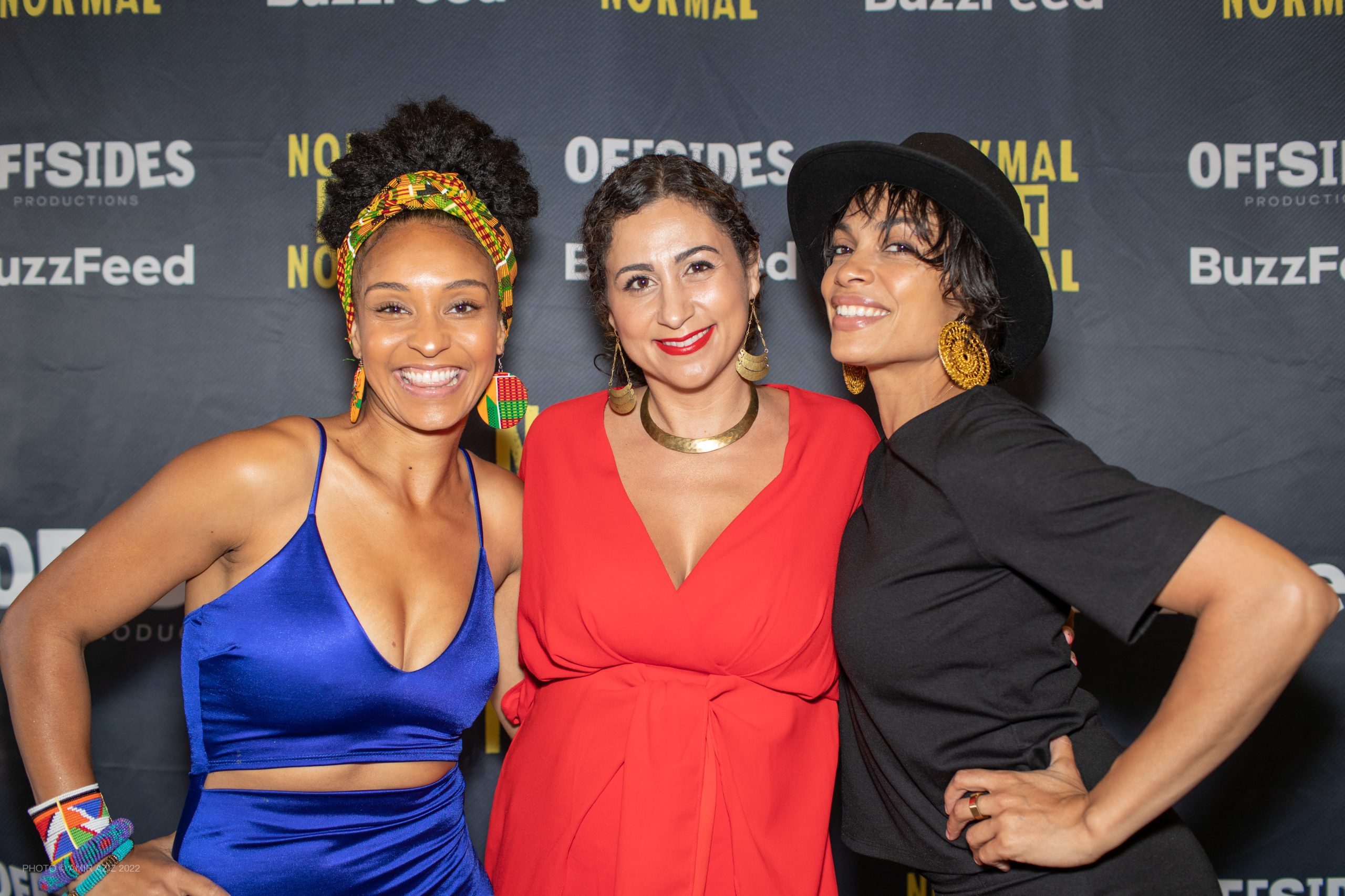 An African American woman wearing a royal blue dress, a Palestinian American woman wearing a bright red dress, and an African American mixed race woman wearing black fedora and black dress pose in front of a black backdrop