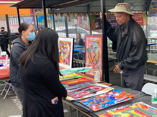 a blind Black man wearing black leather jacket stands behind table that is full of his colorful artwork while he's talking to a guest