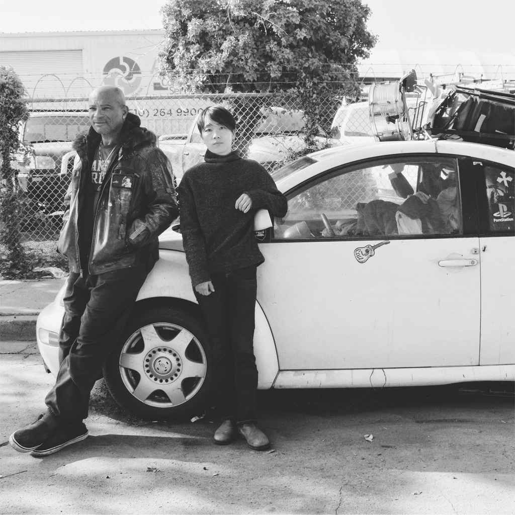 A black and white image with an African American man on the left and Asian American woman with short hair on the right standing in front of a car containing a person's belongings