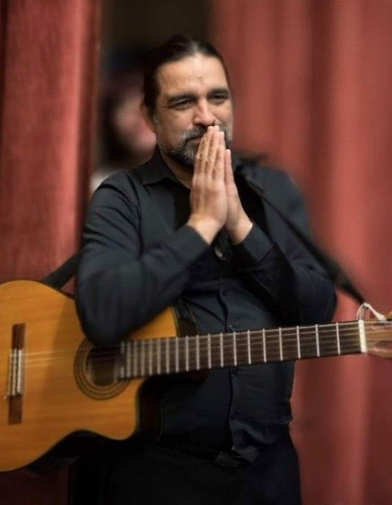 A Latino man holds guitar around him and his hands are in prayer form in front of his face as he is smiling