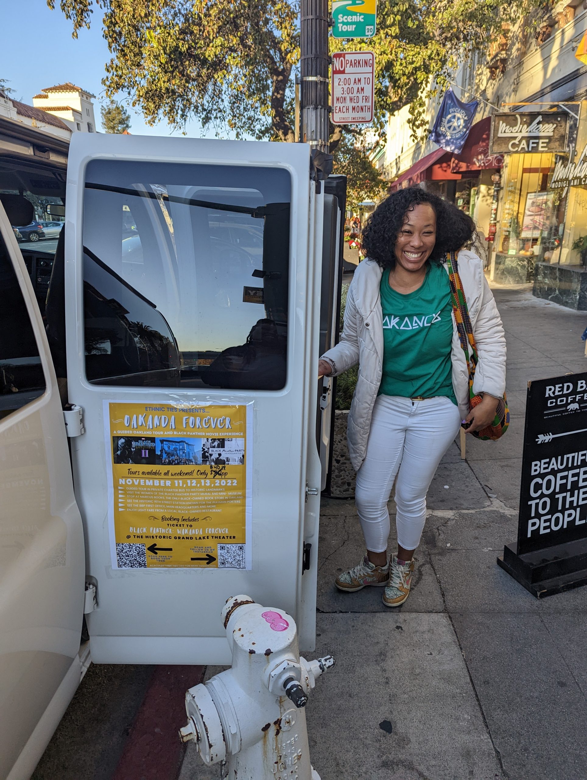 A Black and Chinese woman wearing white jacket and green top smiles next to big van.