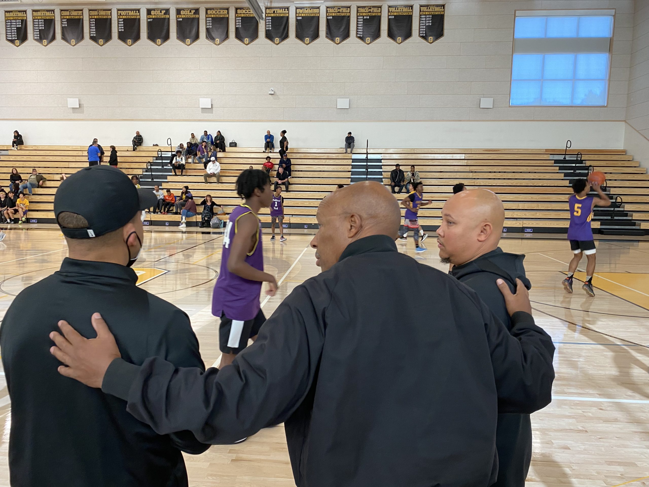 Three male coaches who are friends talk near each other while a scrimmage is going on. The man in the middle has his arms on the other two mens' shoulders
