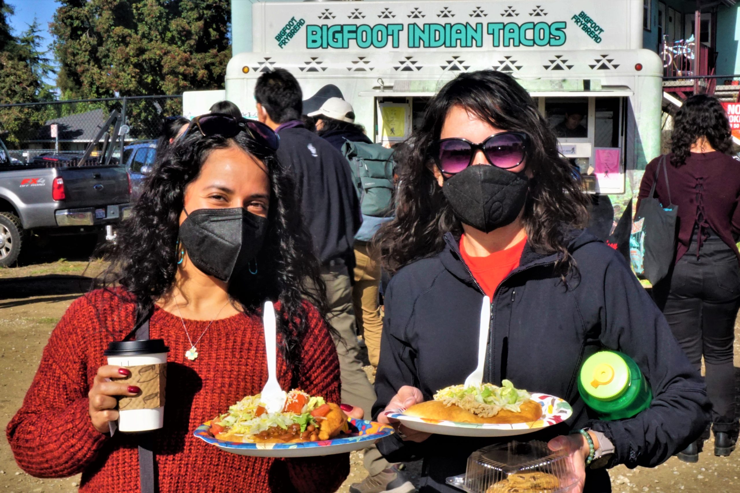 Two women wearing masks hold up plates of fry bread tacos