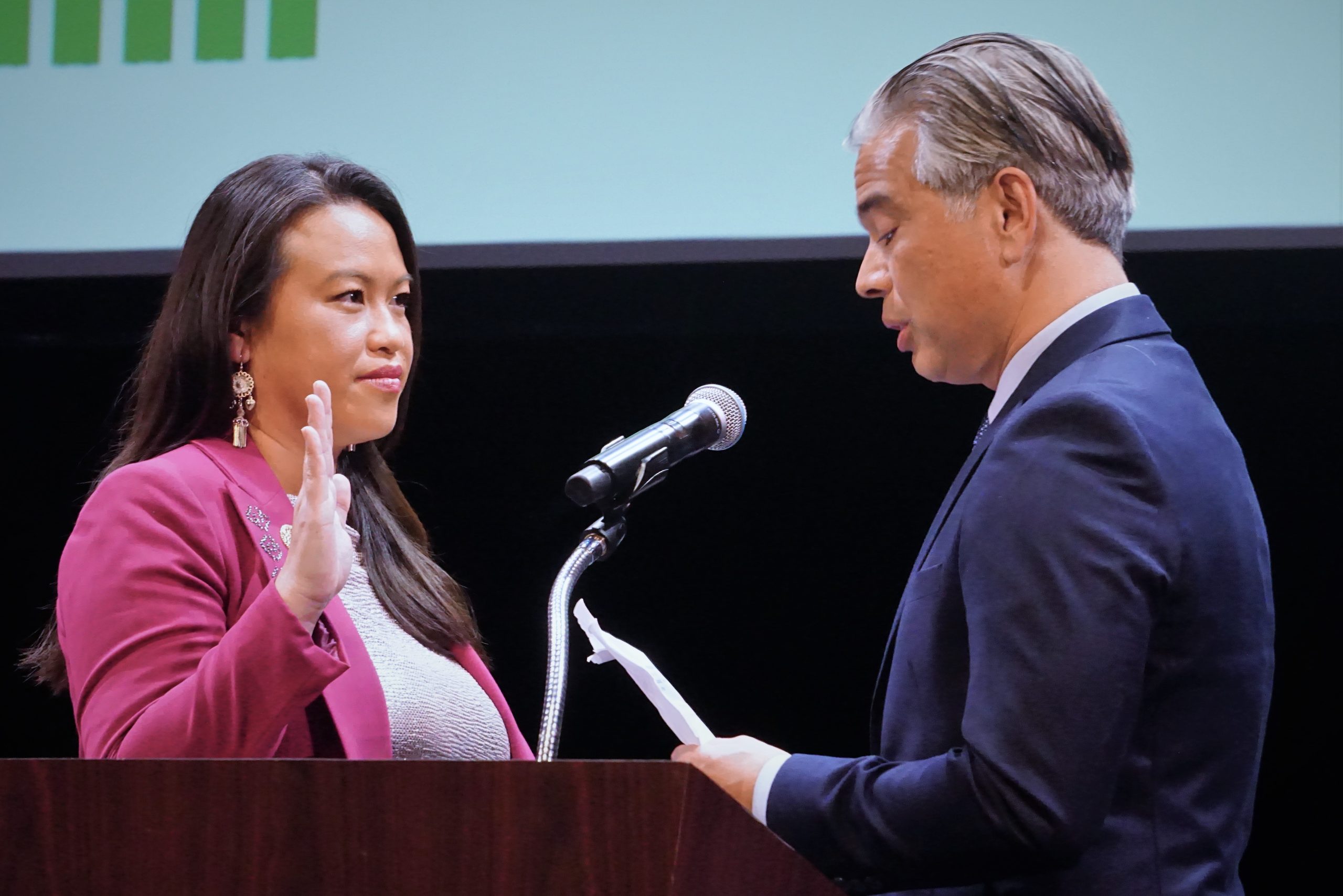 A Hmong American woman wearing purple blazer has her hand up in oath while a Filipino American man speaks by podium mic.