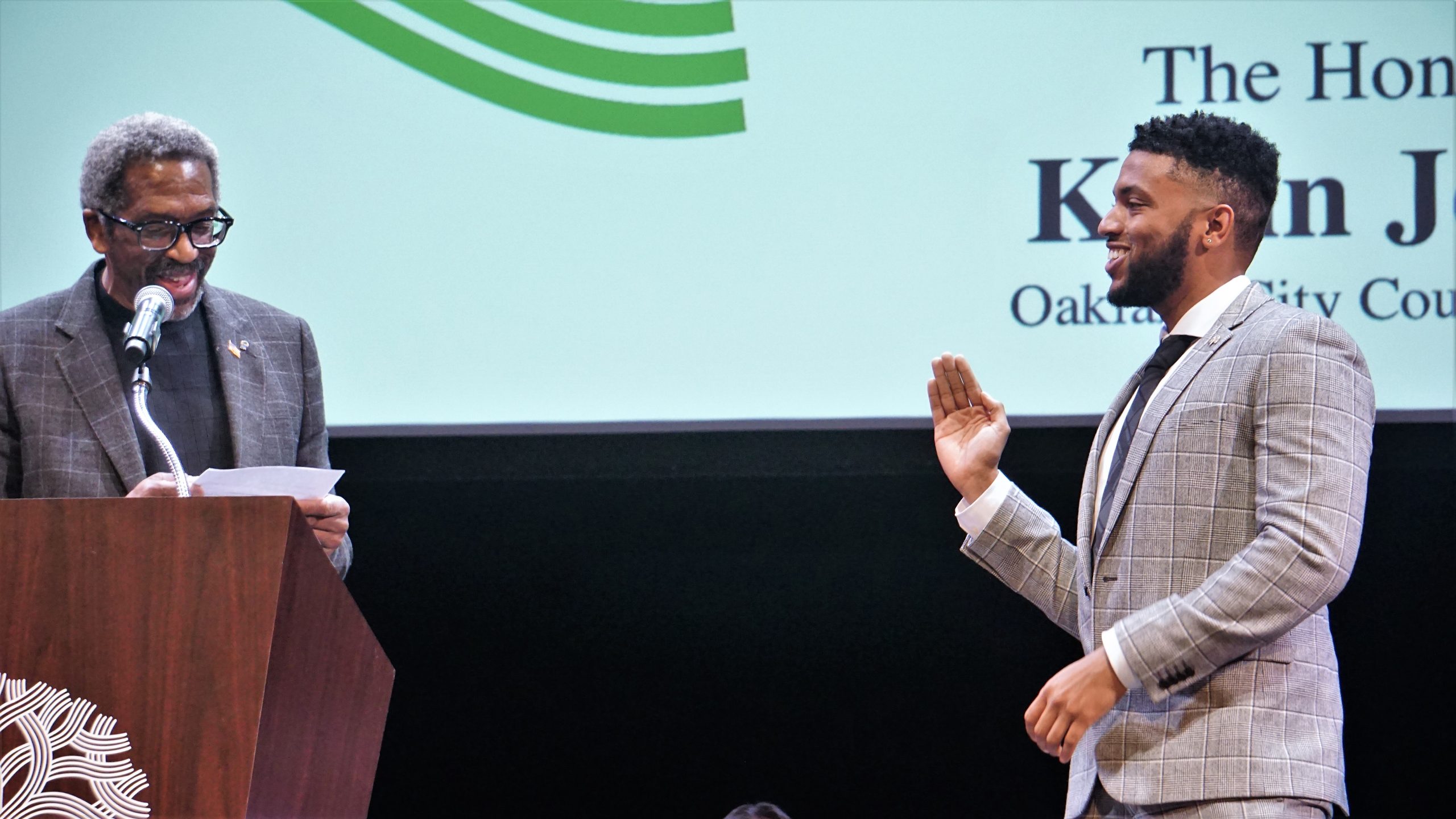 A Black man speaks behind a podium while a younger Black man is sworn in as a city Councilmember.