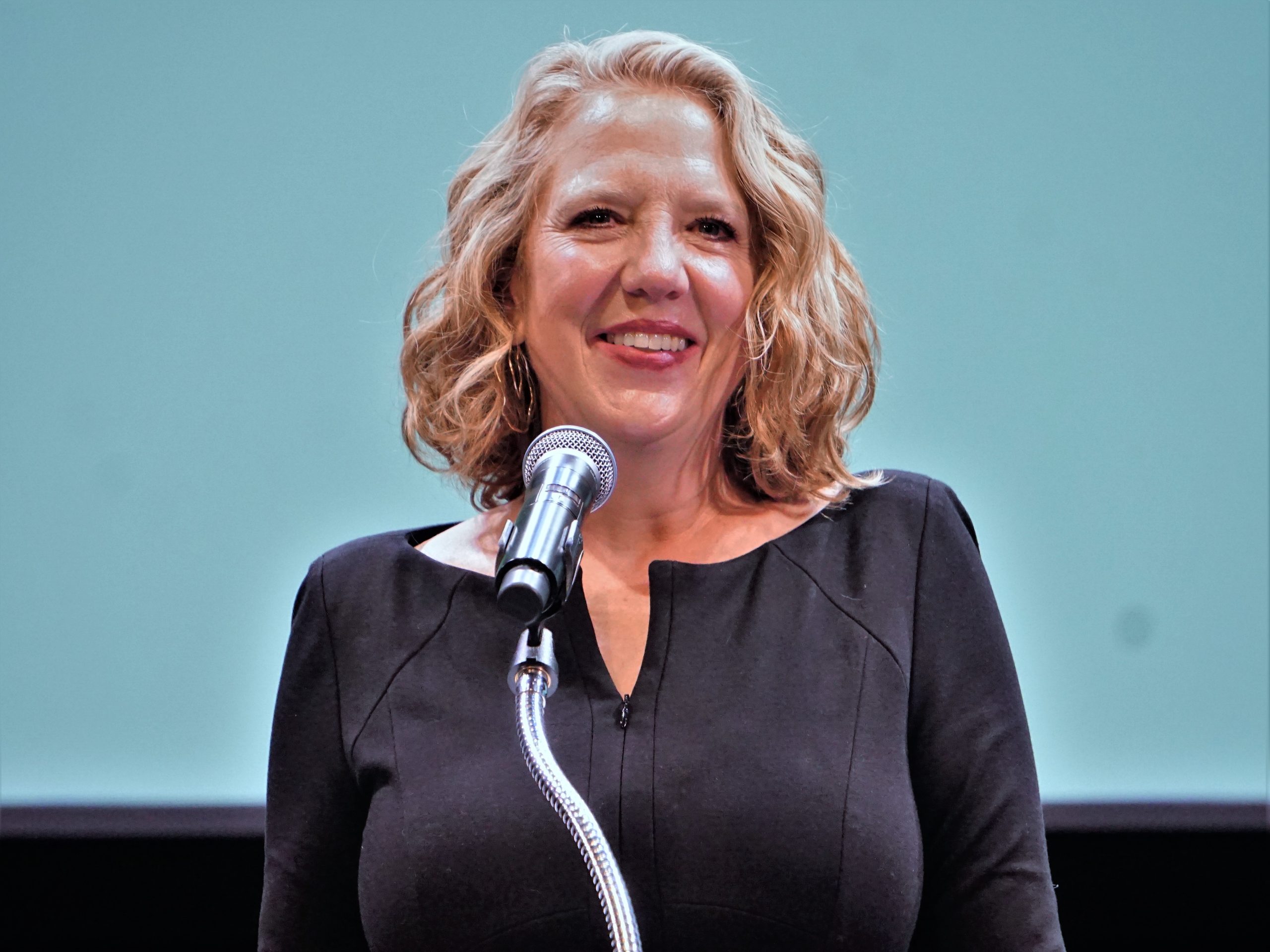 A woman with short curly blond hair stands behind a podium on stage.