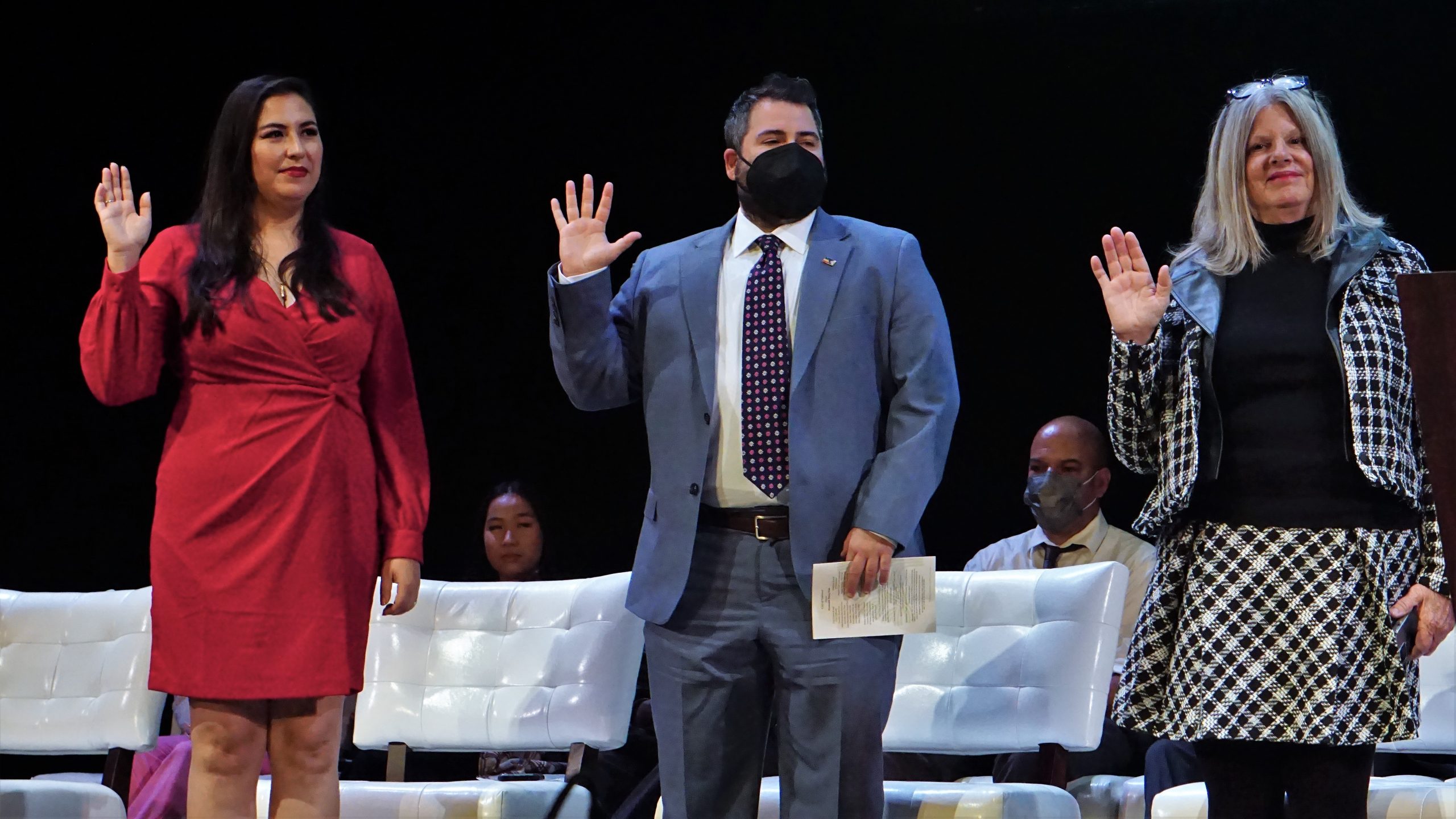 A Bolivian American woman, a trans man and a white woman with grey hair stand on stage in formal wear with their palms facing up