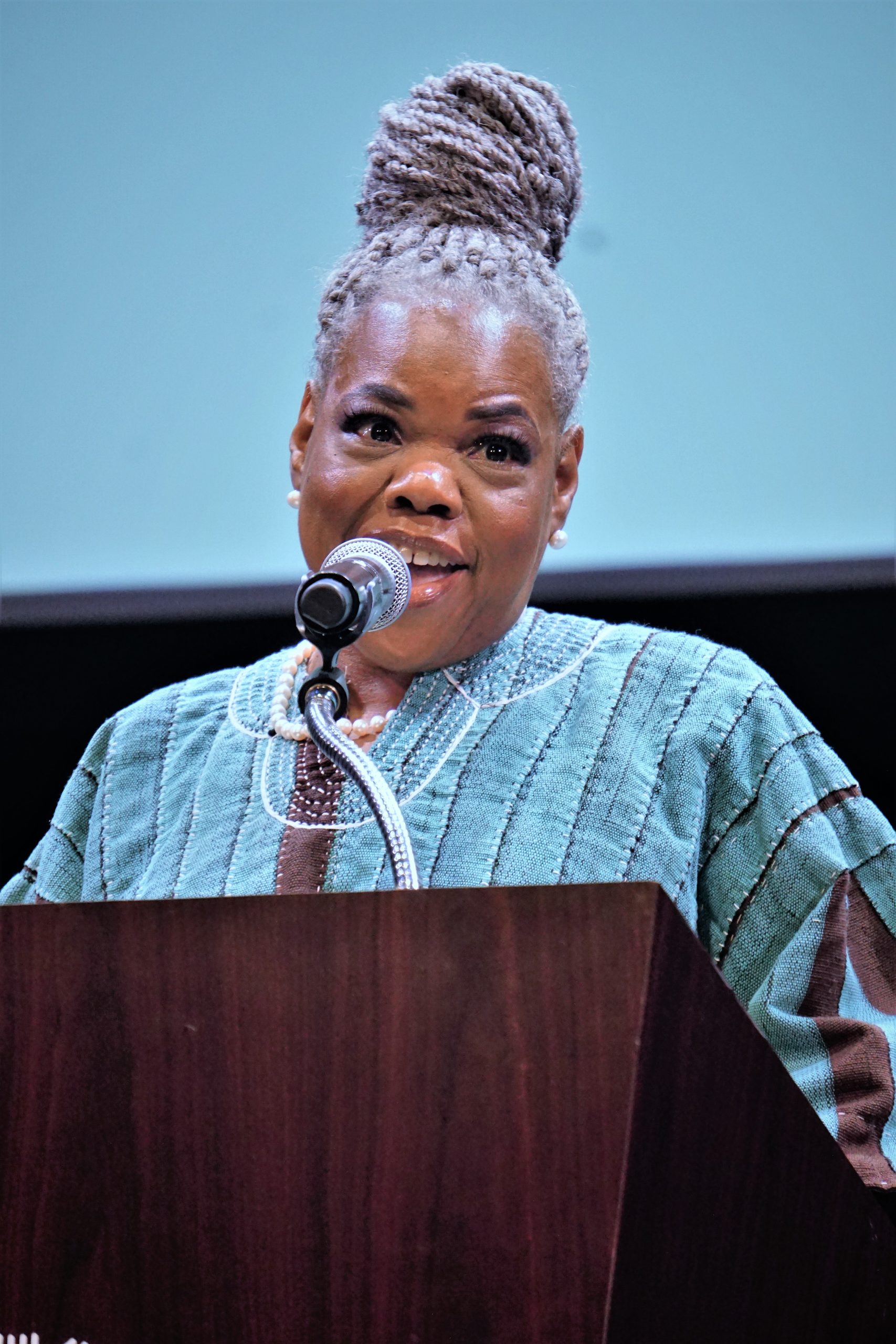 A Black woman with grey braided hair that's in a bun speaks behind podium