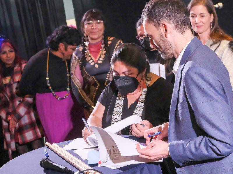 An Ohlone Indigenous woman with long hair wearing a face mask signs and agreement at a public event