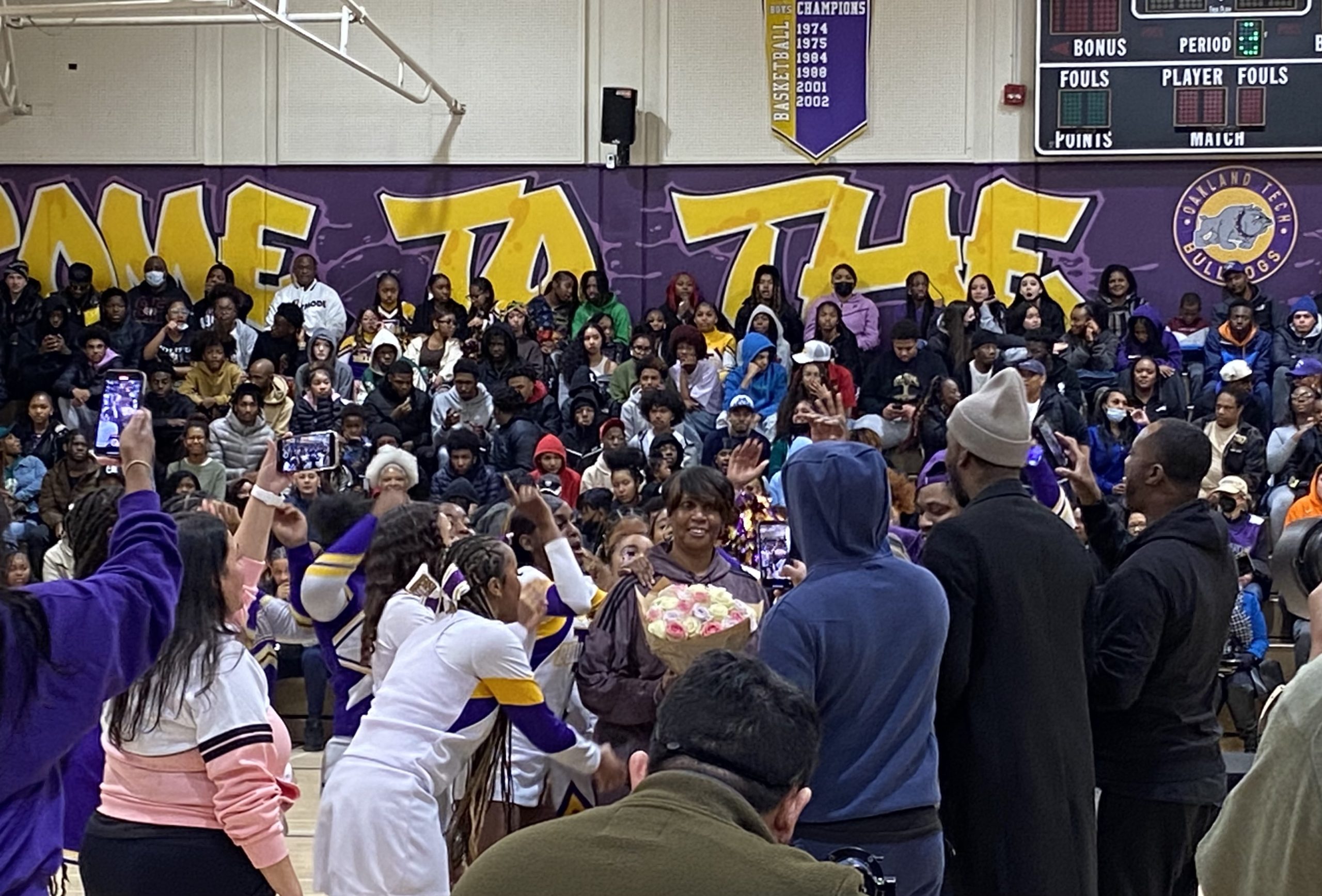 An African American woman is given a big bouquet or roses with many students and adults cheering her on