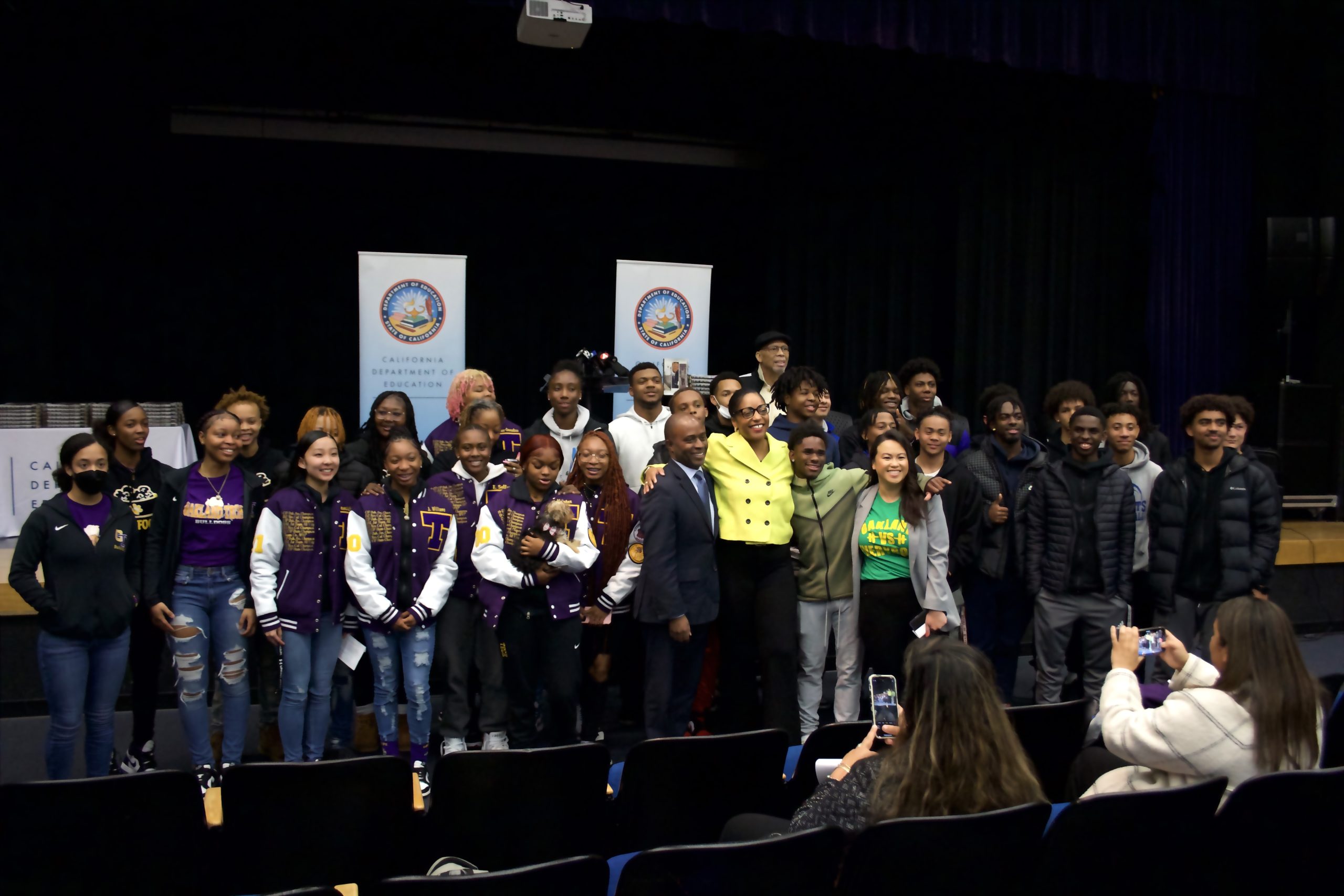 A large group of diverse people and high school basketball players pose for a photo inside an auditorium.