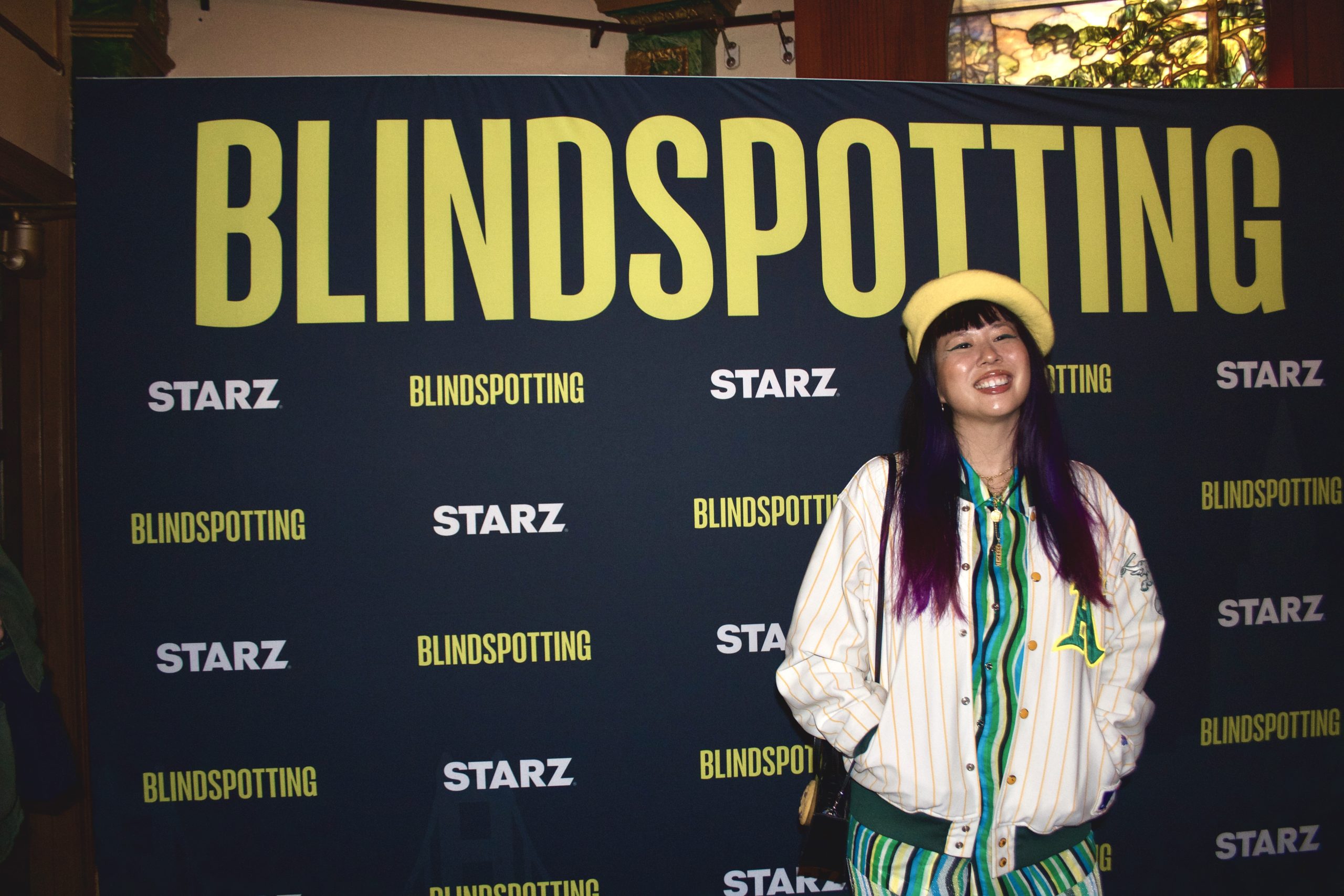 A Taiwanese American woman wearing a yellow hat and green dress smiles at camera while standing in front of a backdrop that says "Blindspotting."