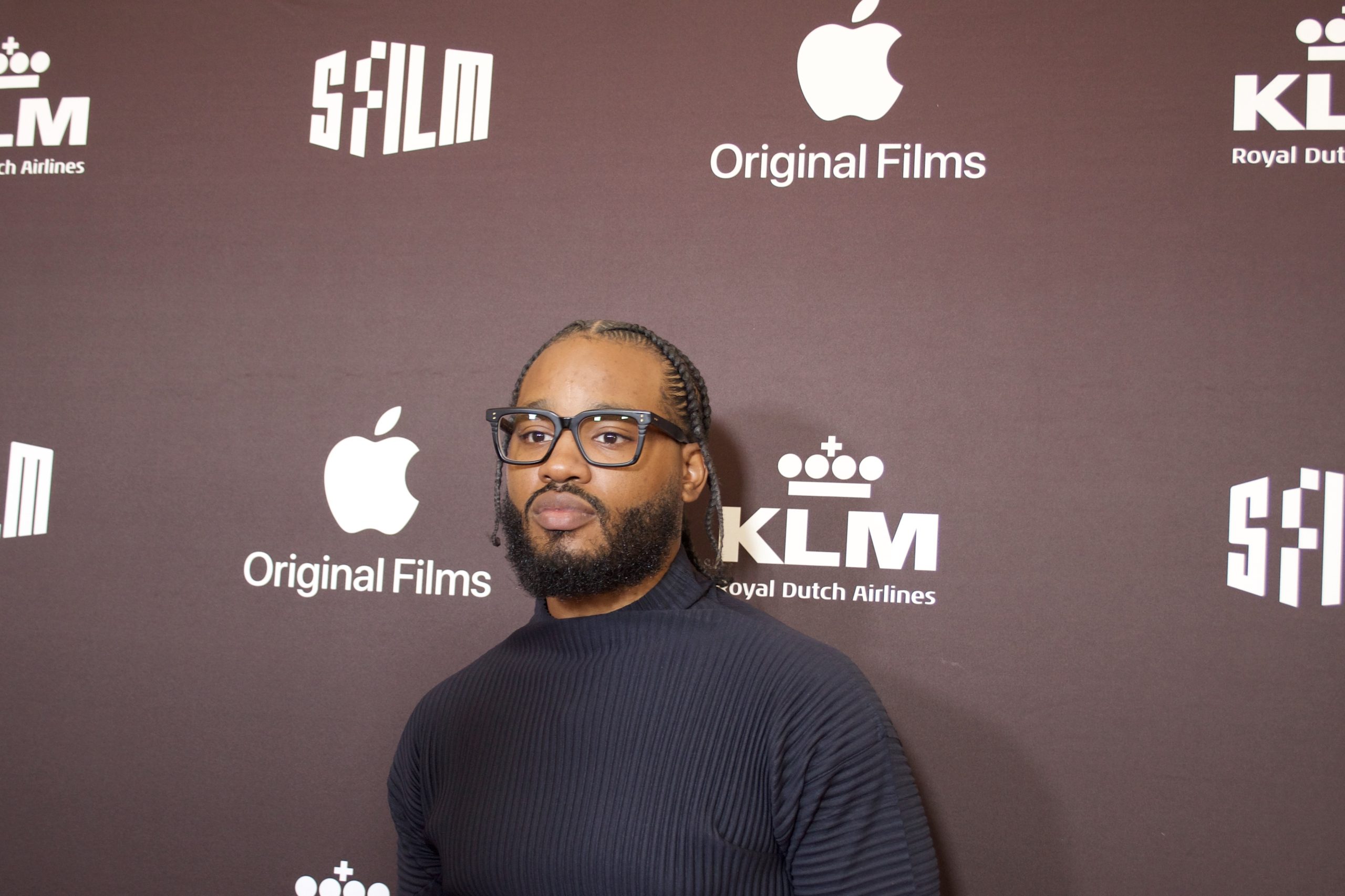 An African American Man with black rimmed glasses stands in front of a brown background for a photo