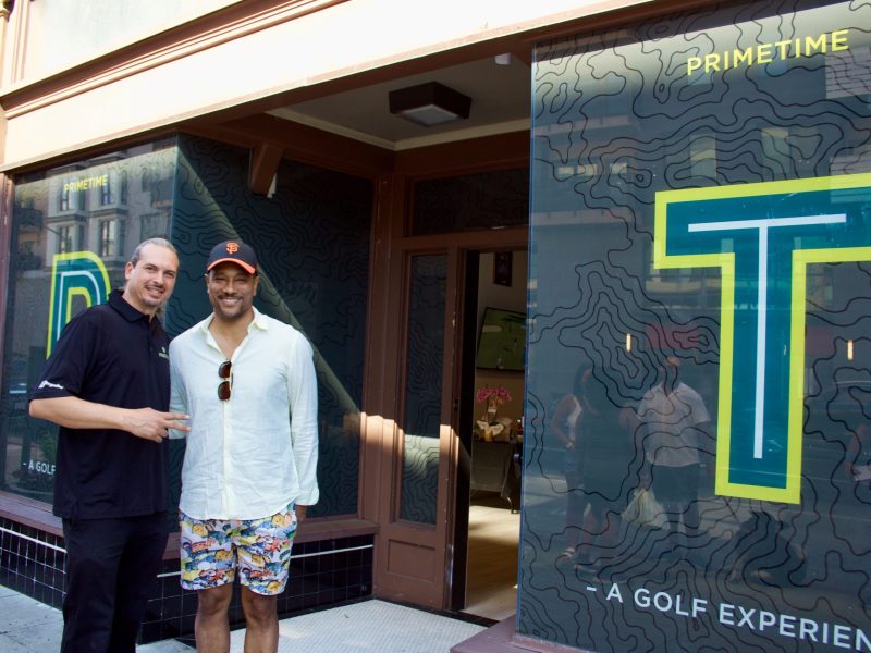 A man with a low pony tail and polo shirt stands next to an African American man in a white shirt in front of a shop that has the letter P and T in large lettering