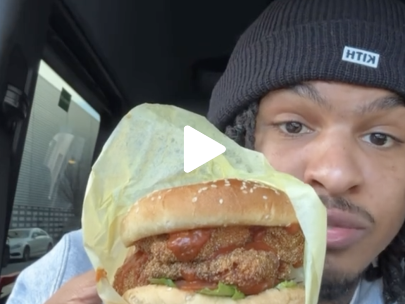 A man sits inside a car and shows the camera a fried fish sandwich