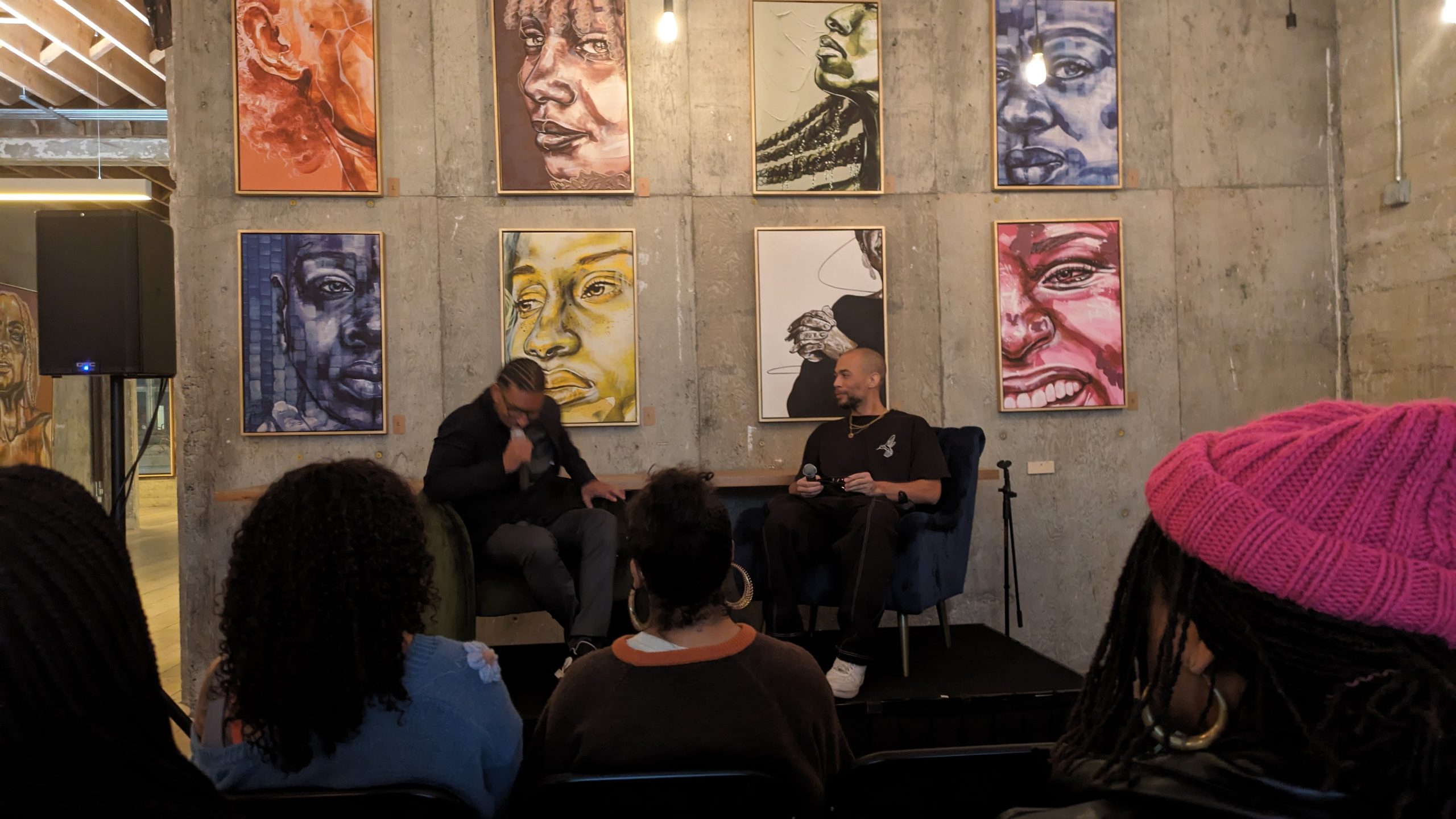 Two Black men sit in front of concrete wall filled with paintings speaking as part of a panel