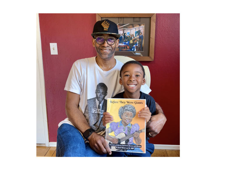 A Black father wearing baseball cap holds a coloring book with his son