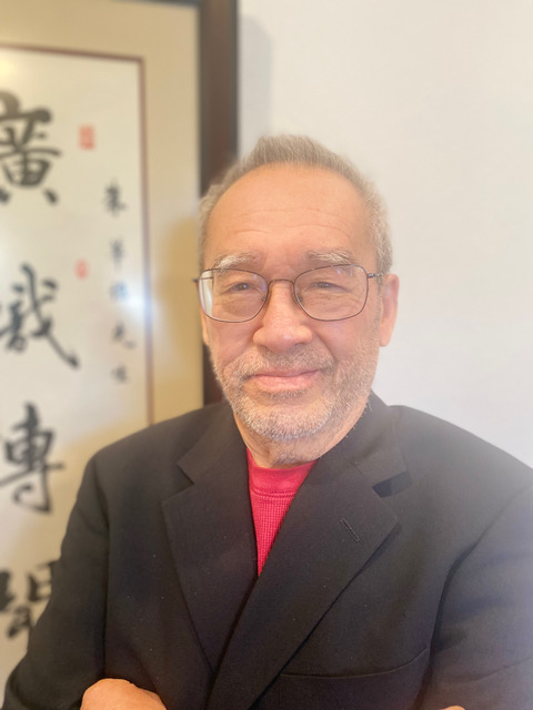 An elder Chinese American man stands for portrait in front of a Chinese painting with Chinese characters