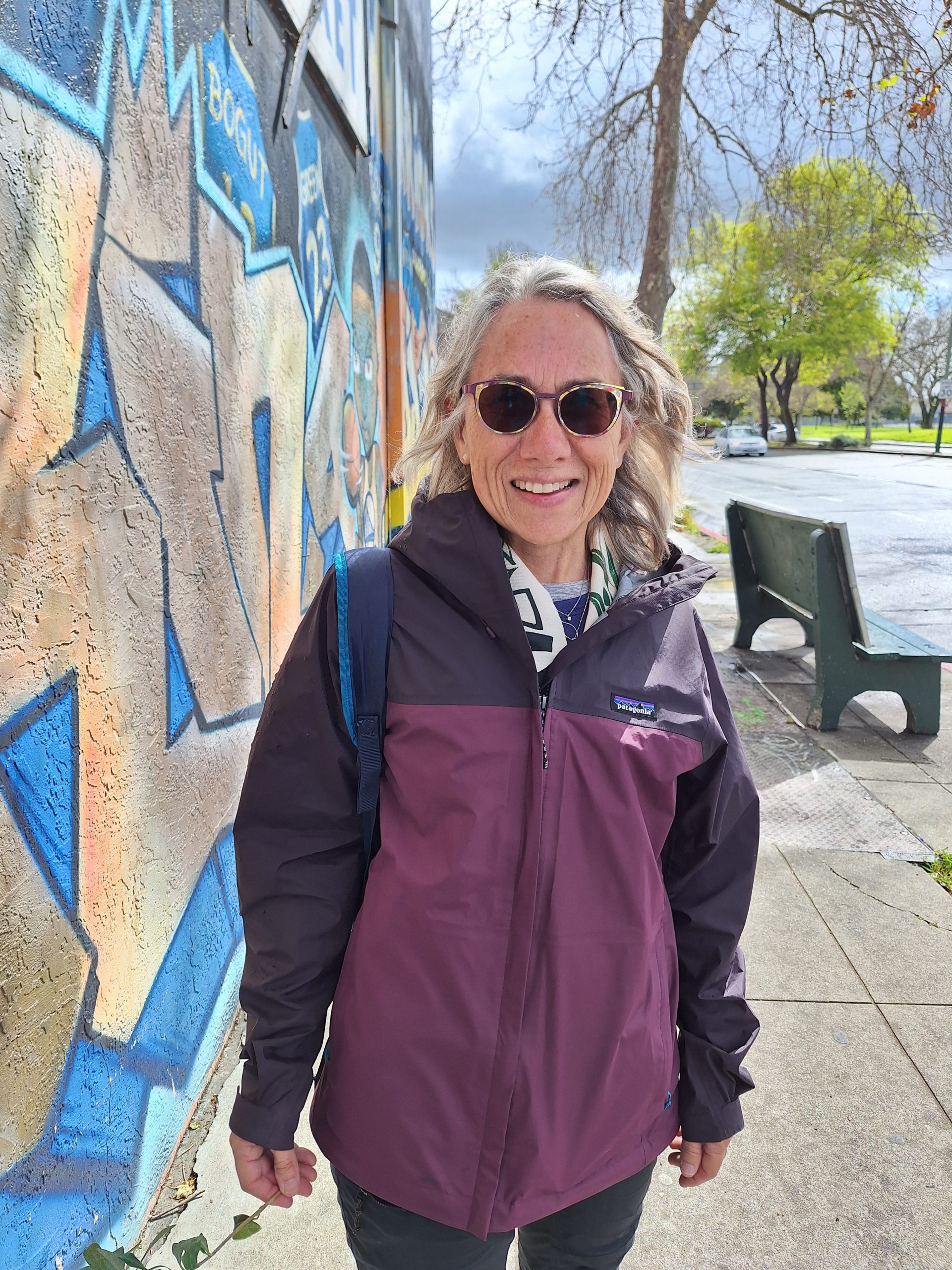A woman with grey hair wearing a purple jacket and sunglasses poses for camera during a spell of sunshine