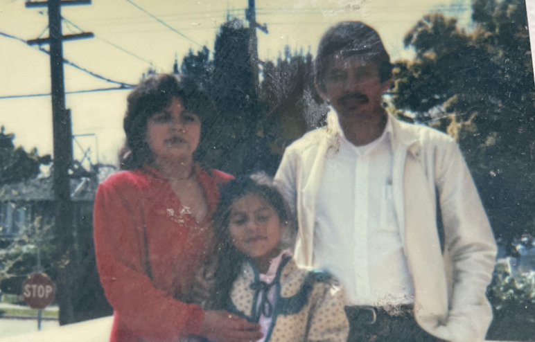 A Latina mom wearing a red suit dress, a young girl, and her father stand in a photo outdoors