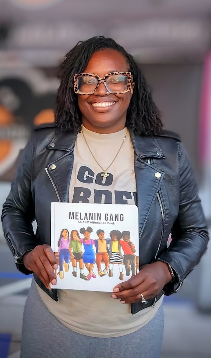 An African American woman wearing glasses holds up a children's book titled "Melanin Gang"